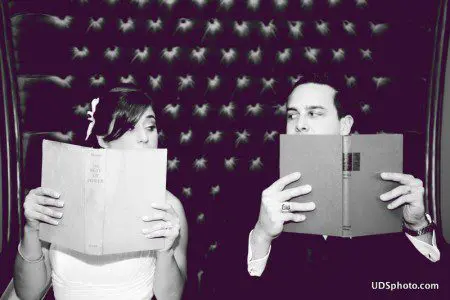 A bride and groom sit side by side, each holding up a book in front of their faces, against a patterned backdrop.