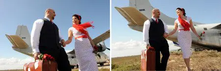 A couple in vintage attire holding hands and suitcases, walking happily near a propeller plane under a clear sky.