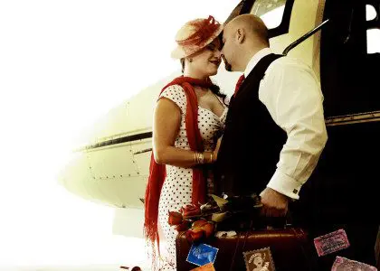 A couple in vintage attire sharing a kiss beside a helicopter, surrounded by luggage, with a focus on romantic and nostalgic aesthetics.