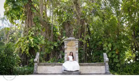Girl in white dress sits on stone bench in lush greenery.