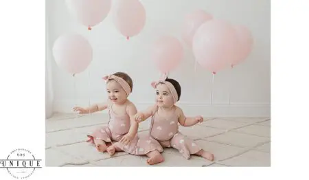 Twin babies in pink outfits with balloons.
