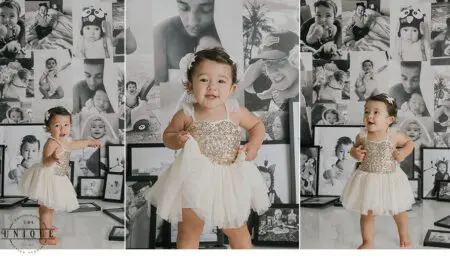 Baby girl in a white dress in front of a photo wall.